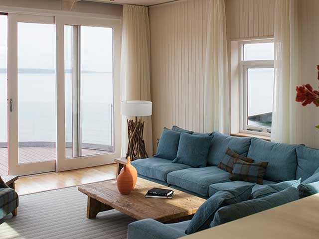 Living area of Grand Designs lifeboat station with deep blue corner sofa, wooden coffee table and sliding clear double door overlooking the sea
