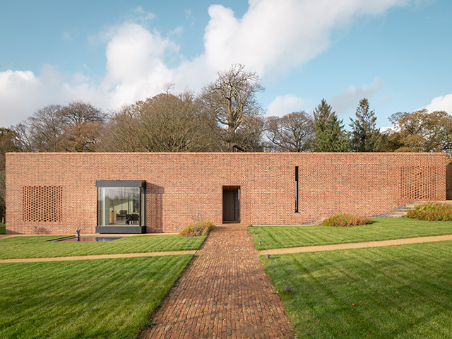 Exterior of a simple single storey house built in red brick that stands out against the green landscape