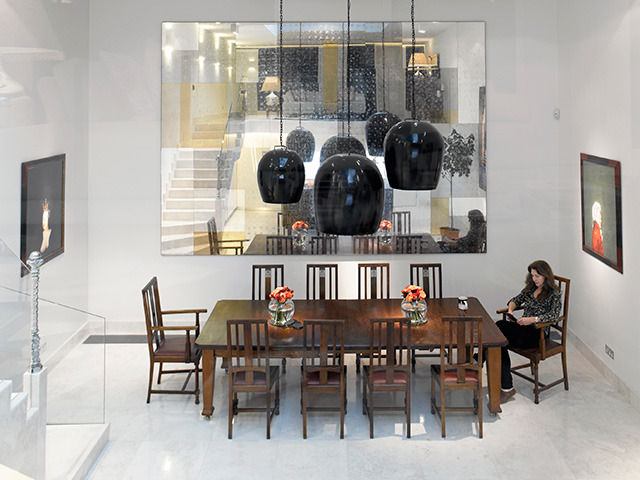 The double-height dining room has a hanging black light fitting, white interiors and wooden furniture across the 10 seater table.