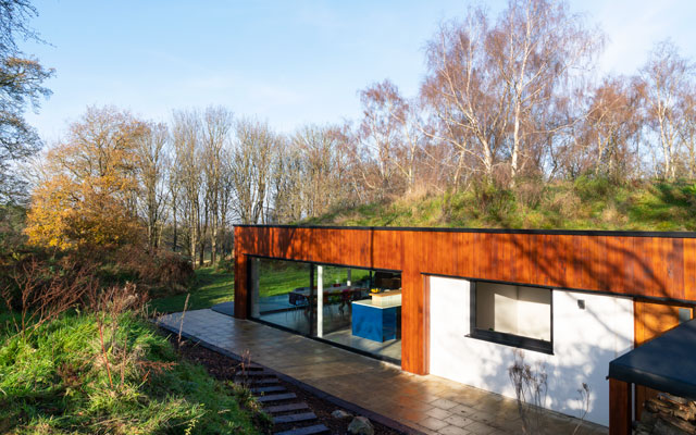 Side view of the self-heating house exterior showing the red cedar cladding and paved deck