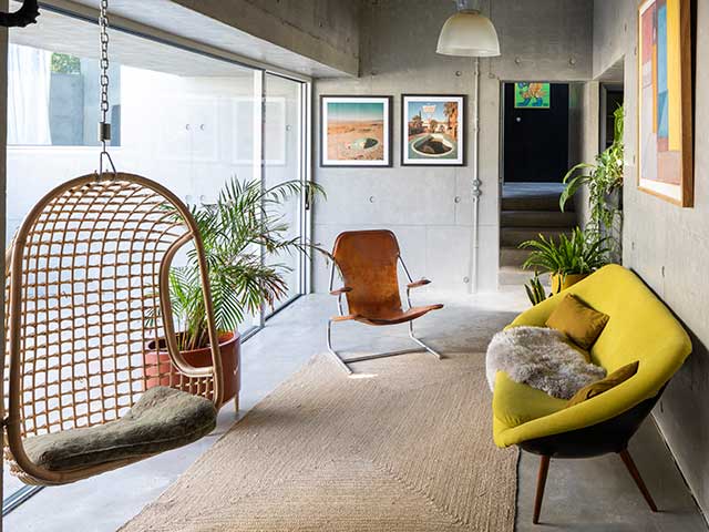 The living space in the Grand Designs concrete house with wooden hanging chair and yellow sofa overlooking sliding doors into garden