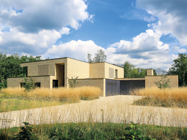 Large contemporary yellow brick home with paved drive surrounded by woodland