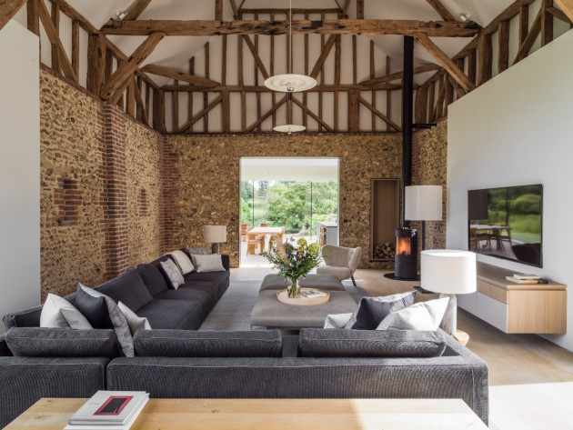 interior of barn conversion with stone walls exposed beam roof and grey contemporary modular sofas wooden floors and cylindrical wood burner