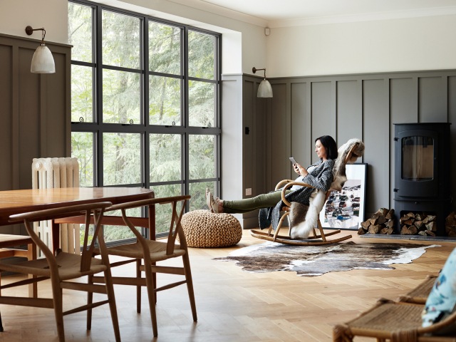 Wood-panelled contemporary space with log burner, woman in rocking chair and industrial-style black Sheerline classic aluminium windows