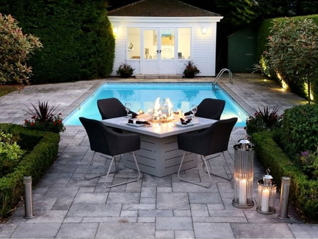 Square table outside furniture beside pool with floor standing candles and white annexe