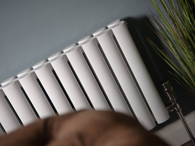 white oval tube radiator against a grey wall with leather sofa and house plants