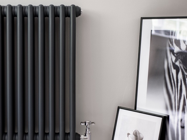 dark grey radiator with oval fins and chrome valves against a pale grey wall with wall art