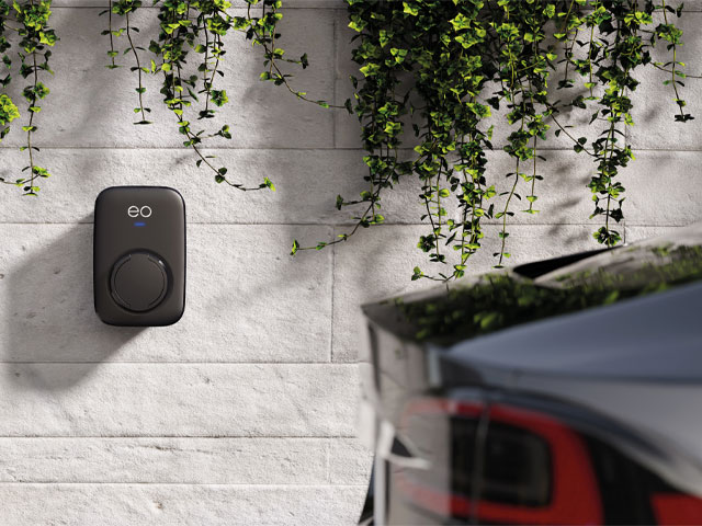black designer home ev charger against a white brick wall with hanging plants and black car