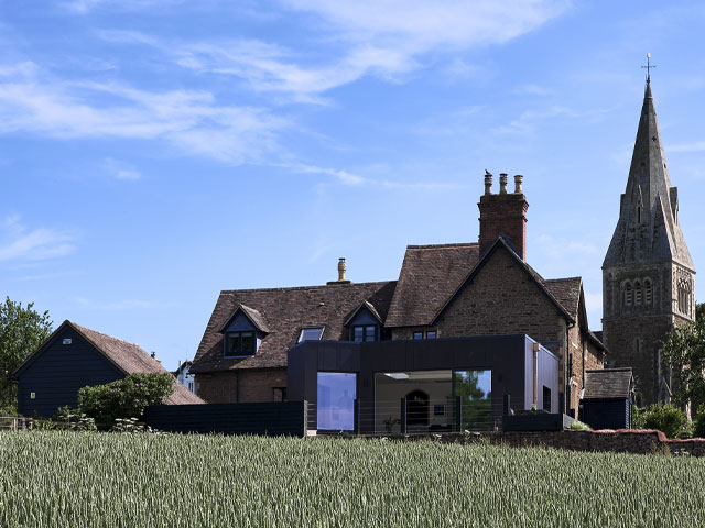 large stone cottage in the countryside in front of a church spire with a timber-clad kitchen addition to the rear 
