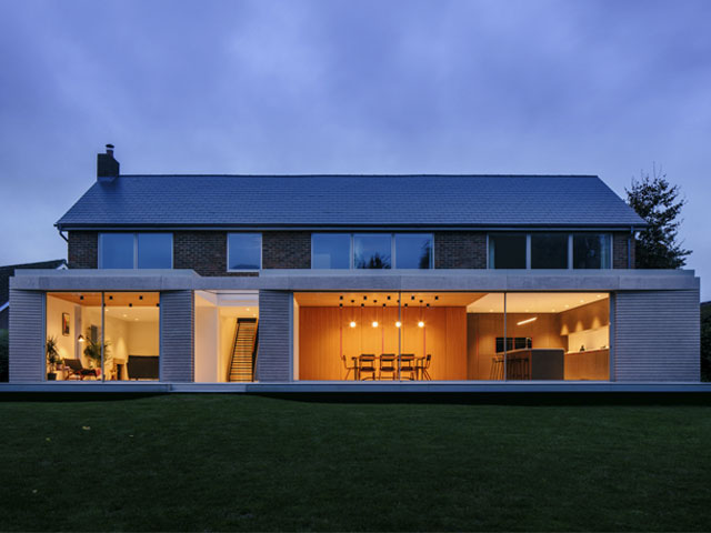 large rear extension to a 60s brick built house made of concrete and glass photographed at dusk with the lights on