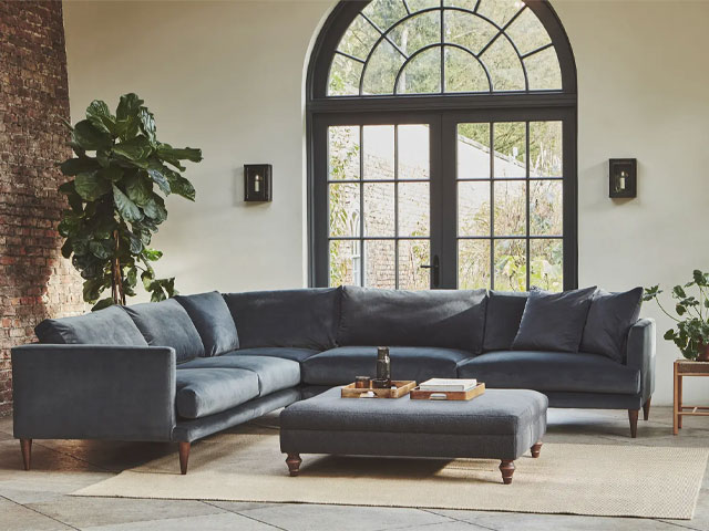 blue velvet corner sofa with scatterback cushions and teak legs in a a living room with exposed brick and a large arched window
