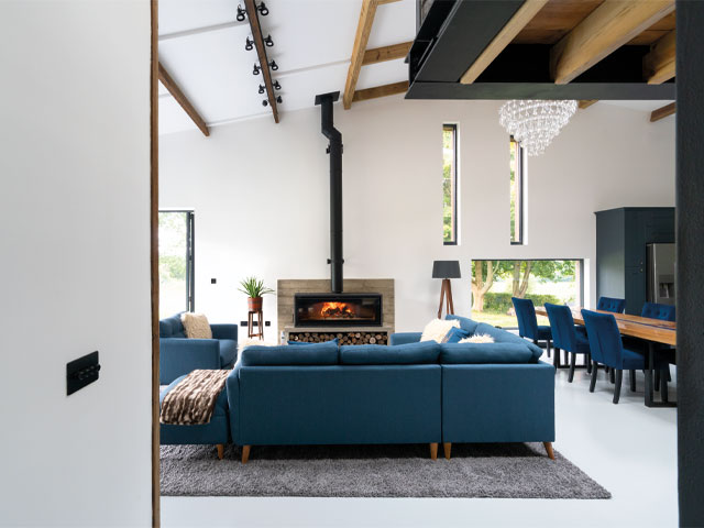  Grand Designs renovations. Double height ceiling large wood burning fire wooden dining table blue chairs large blue l-shaped sofa exposed wooden beams