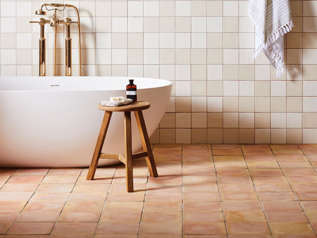 Freestanding white bath with copper taps terracotta tiles wooden chair with shampoo bottle on top 