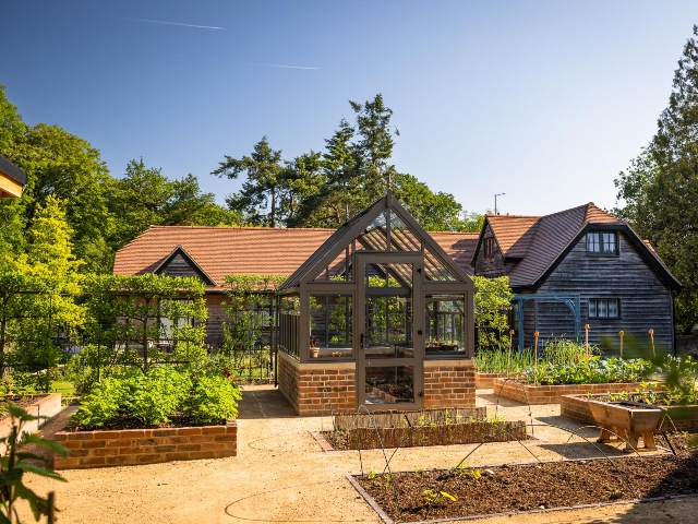 Aluminium greenhouse with brick base and brick planters in foreground of wood pannelled home
