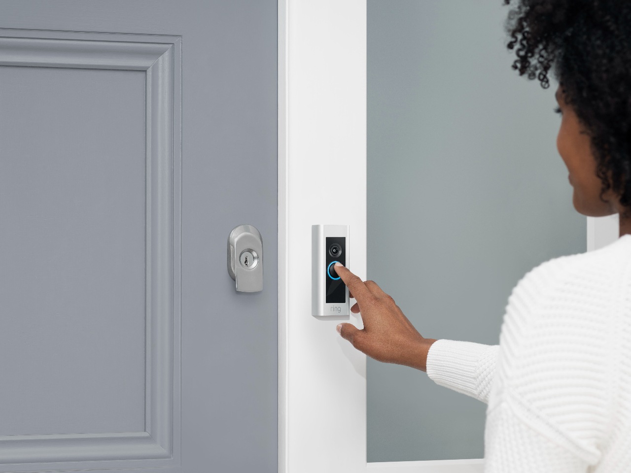 A woman pressing the button on a Ring Video Doorbell Pro, outside a grey door with a white frame