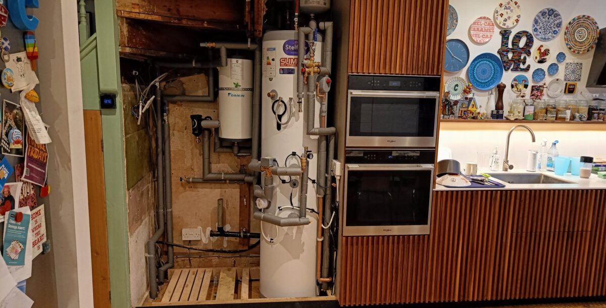 The cylinders and plumbing of an air source heat pump in a tall, walnut kitchen cabinet
